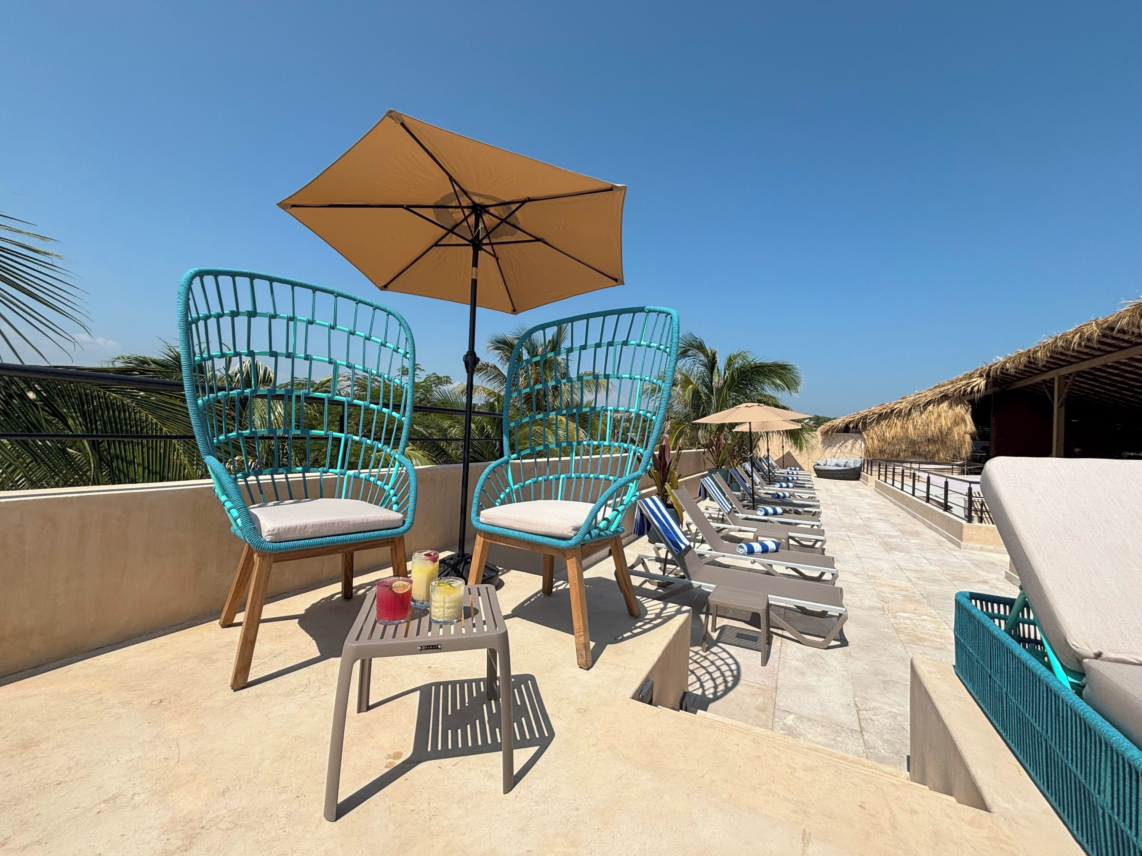 A rooftop terrace with turquoise wicker chairs, a table with drinks, and lounge chairs.