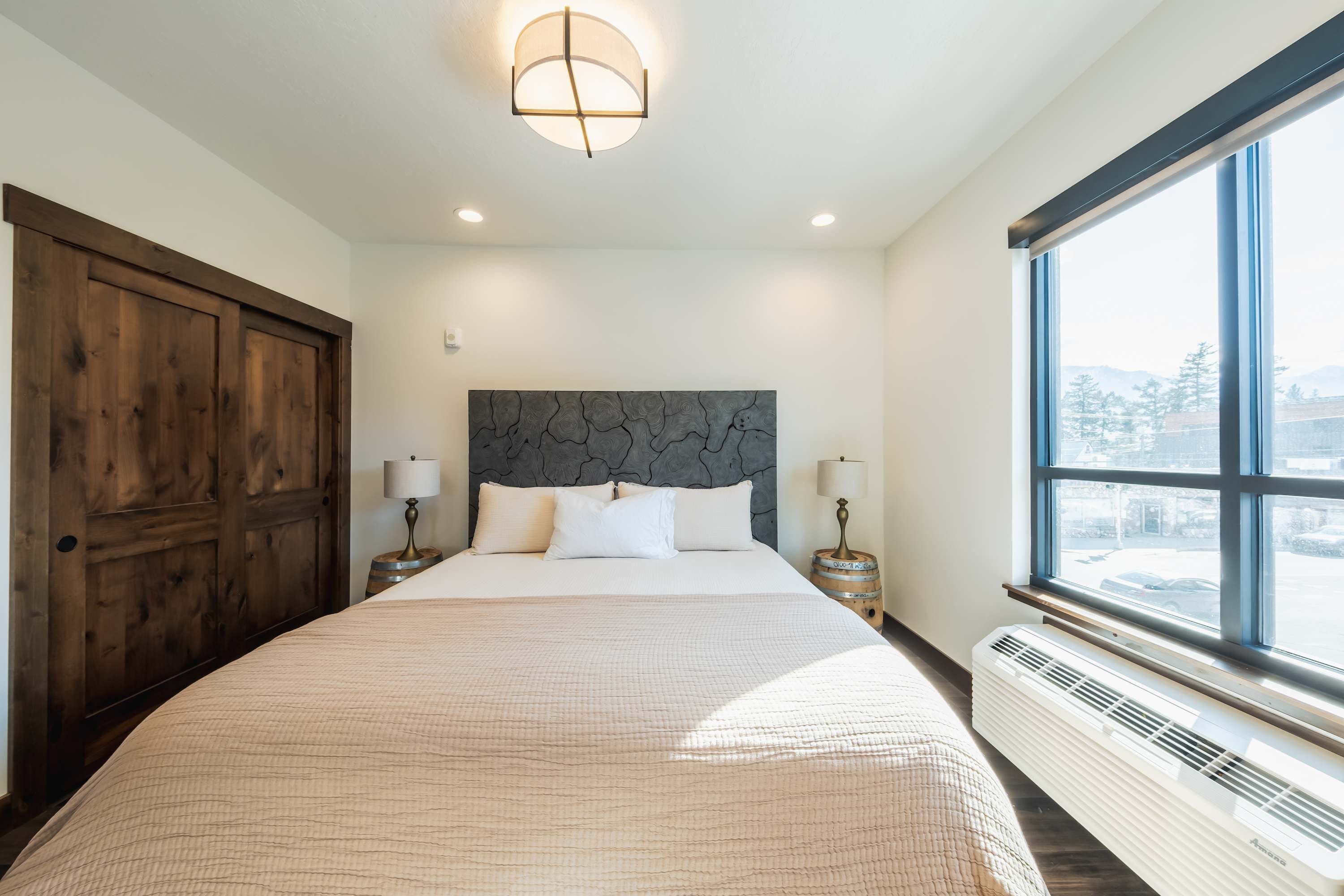 A bedroom with a large bed, wooden wardrobe, bedside lamps, and a view through a large window.