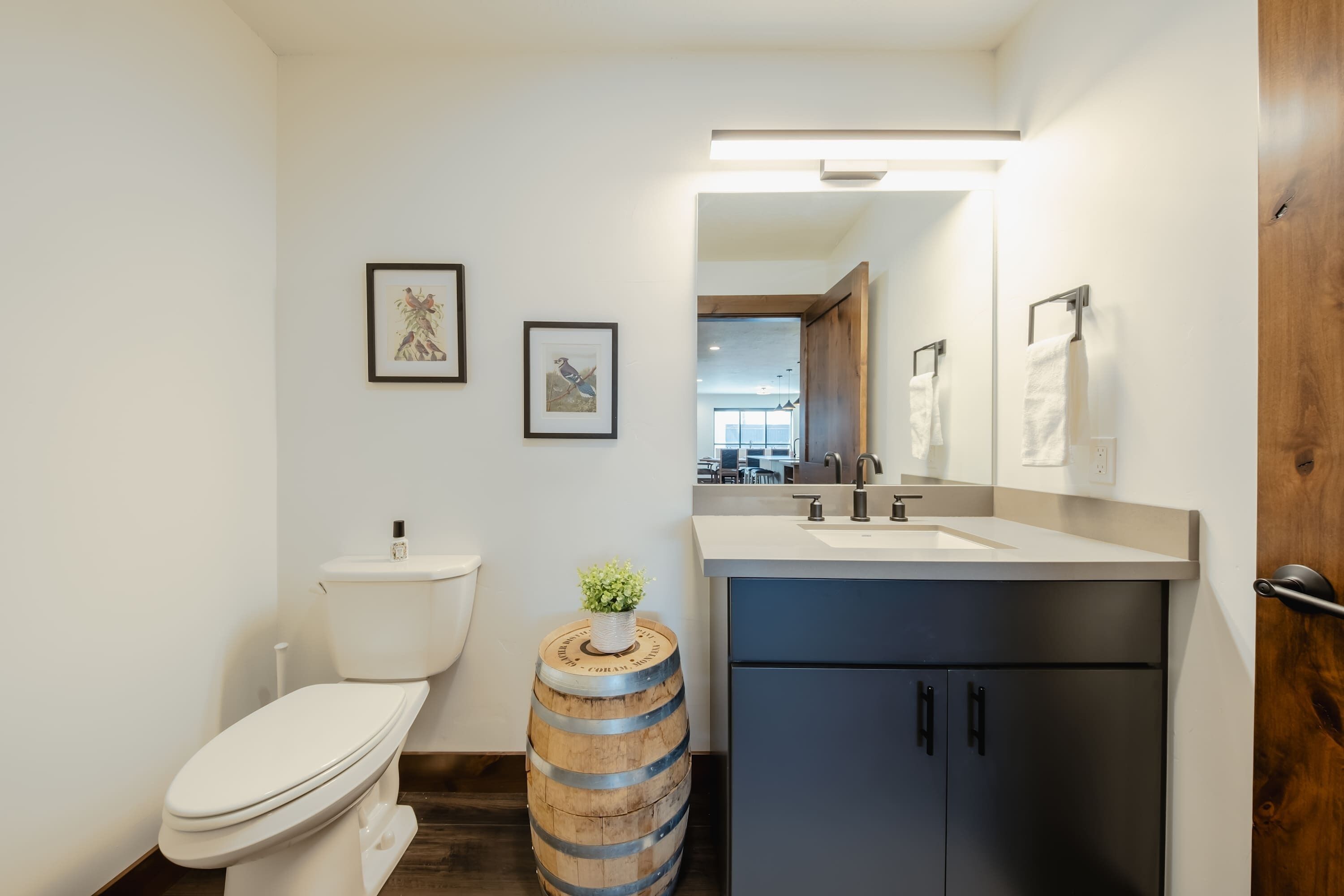 Un bagno moderno con il water, un lavandino e uno specchio. Due quadri appesi alla parete e una piccola pianta su una botte.