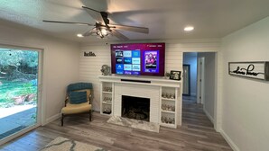 Una sala de estar con chimenea, un televisor instalado que muestra servicios de streaming, un ventilador de techo y vistas al exterior a través de una puerta de cristal.