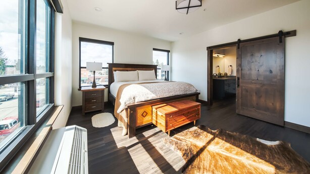 A modern bedroom with a large bed, a wooden bench, a large window, and a sliding door. - Downtown Penthouse Condo For 4! (Columbia Falls)