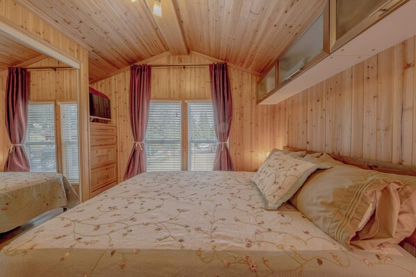 Un dormitorio de cabaña de madera con una cama, dos ventanas con cortinas y un televisor pequeño en la pared.