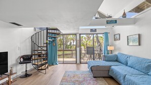 Un salon moderne doté d’un canapé incurvé, d’un escalier en colimaçon et d’une grande porte-fenêtre coulissante donnant sur un espace extérieur avec vue sur des palmiers.
