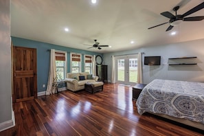 A spacious bedroom with hardwood flooring, a large bed, a ceiling fan, and a television mounted on the wall.