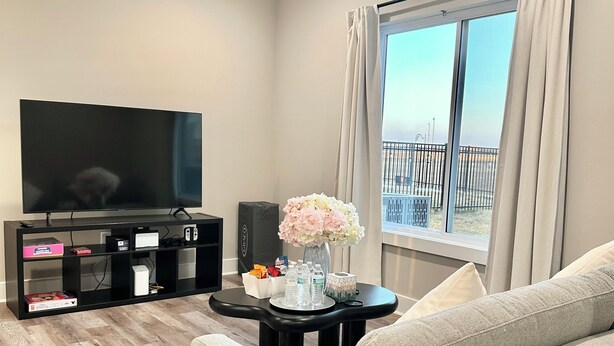 A living room with a flat-screen TV, a black entertainment unit, a grey sofa, a small round coffee table with a vase of flowers, and a large window with curtains.