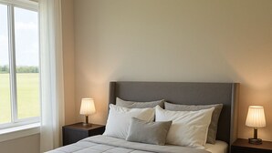 A neatly made bed with a grey headboard, two bedside tables with lamps, and a window with a view of a green field.