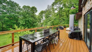 A wooden deck with a glass-top table and chairs, surrounded by lush greenery and a stone building.