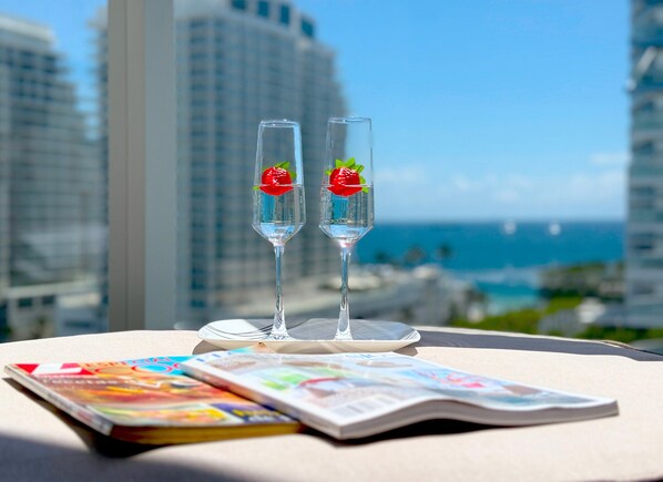 Two champagne flutes with strawberries on a table with magazines, overlooking a cityscape and the ocean.