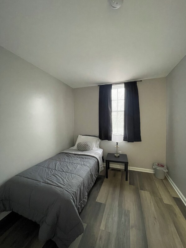 A small, neatly arranged bedroom with a single bed, a nightstand, a lamp, and a window with curtains.