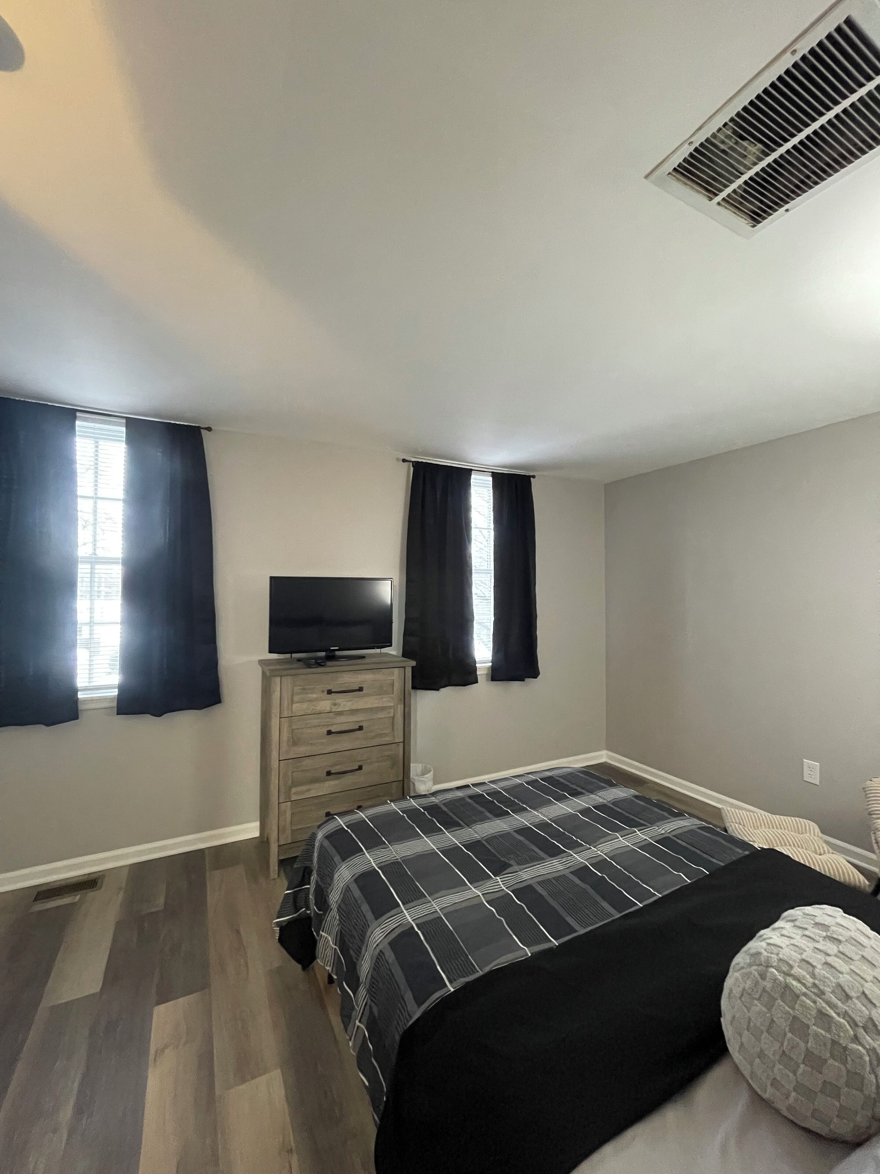 Quarto com cama, cômoda, televisão e duas janelas com cortinas.