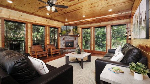 A cozy living room with a fireplace, leather sofas, and large windows offering a view of the forest. - Soak Among the Treetops, Swim Indoors, Shoot Pool & Host Luxe Movie Nights - Whispering Pines Escape (Sevierville)
