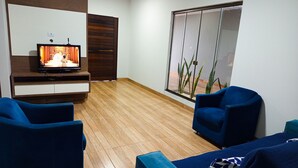 A modern living room with a flat-screen TV, two blue sofas, a wooden floor, and a large window with plants. - Big House Leisure 🌊 (Dourados)