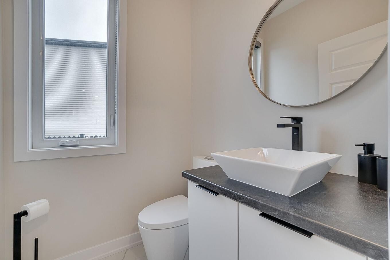Et moderne badeværelse med en hvid vask, en sort bordplade, et toilet og et vindue med persienner.