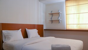 Un letto rifatto con lenzuola bianche e un asciugamano piegato, una testiera in legno e una mensola a parete con un punto luce.