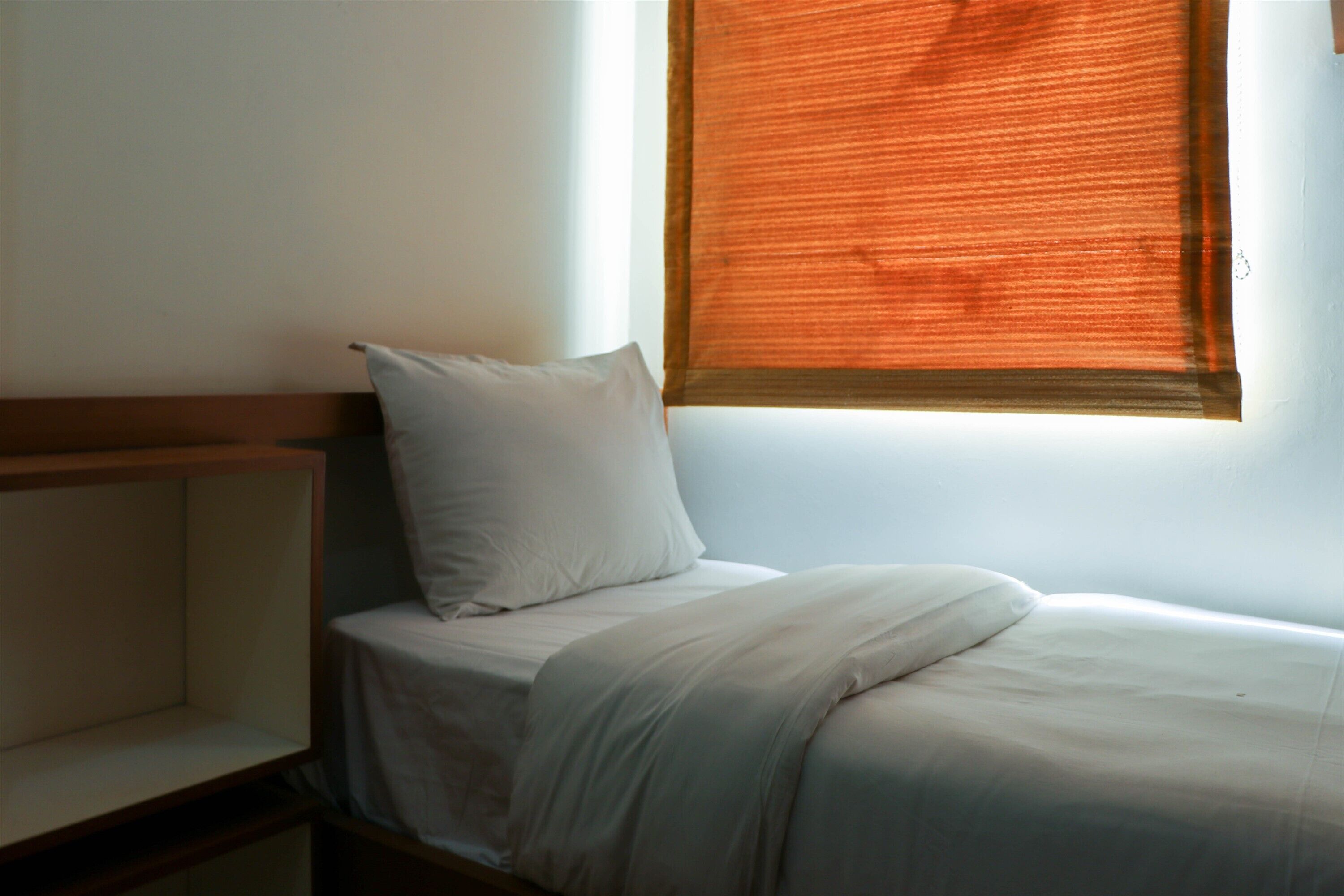 Ein Einzelbett mit weißer Matratze und Kissen, ein Holz-Kopfteil und ein Fenster mit orangefarbenen Jalousien.