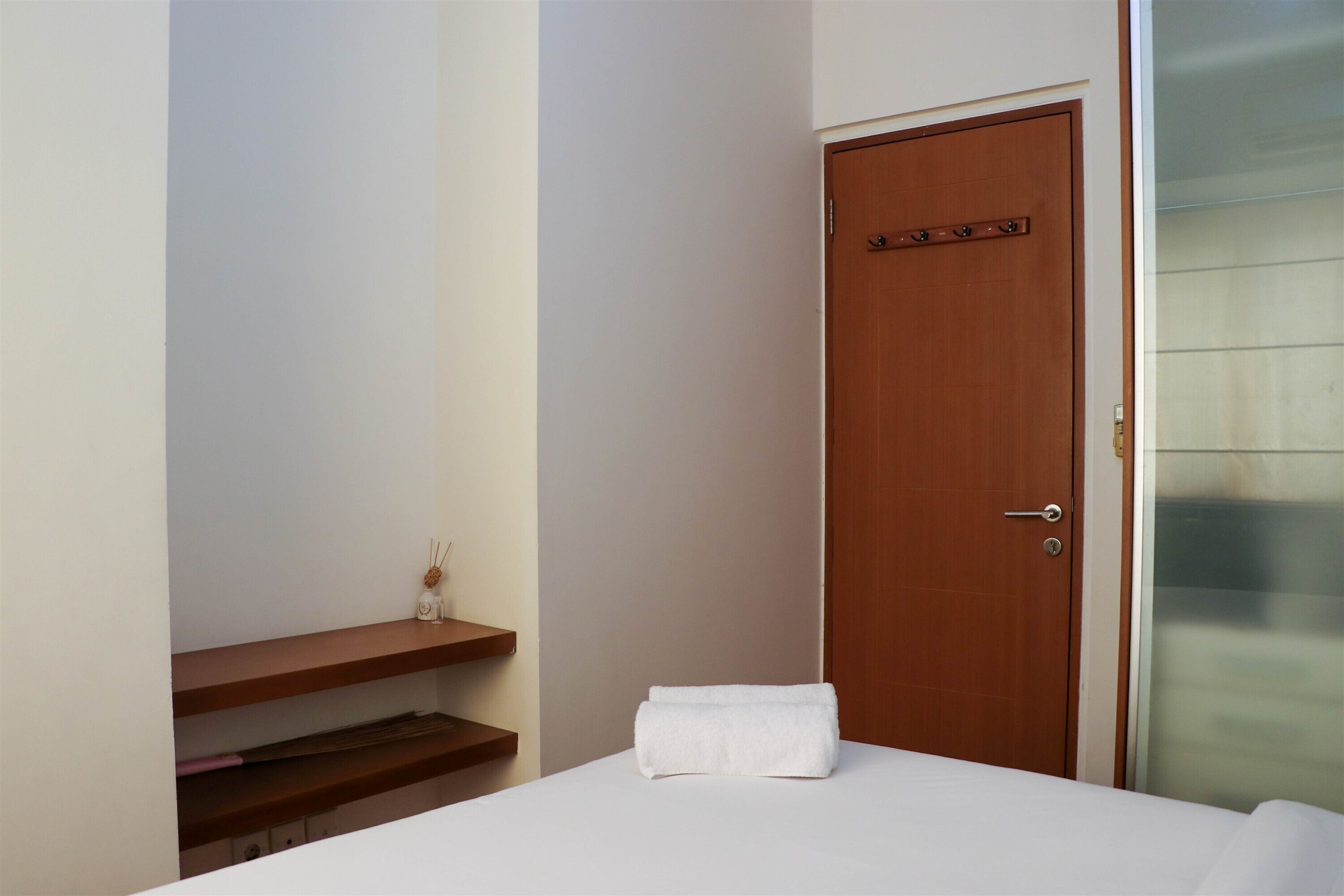 A hotel room with a wooden door, a white bed with a neatly folded white towel, a wooden shelf with a small bottle, and a glass door to the right.