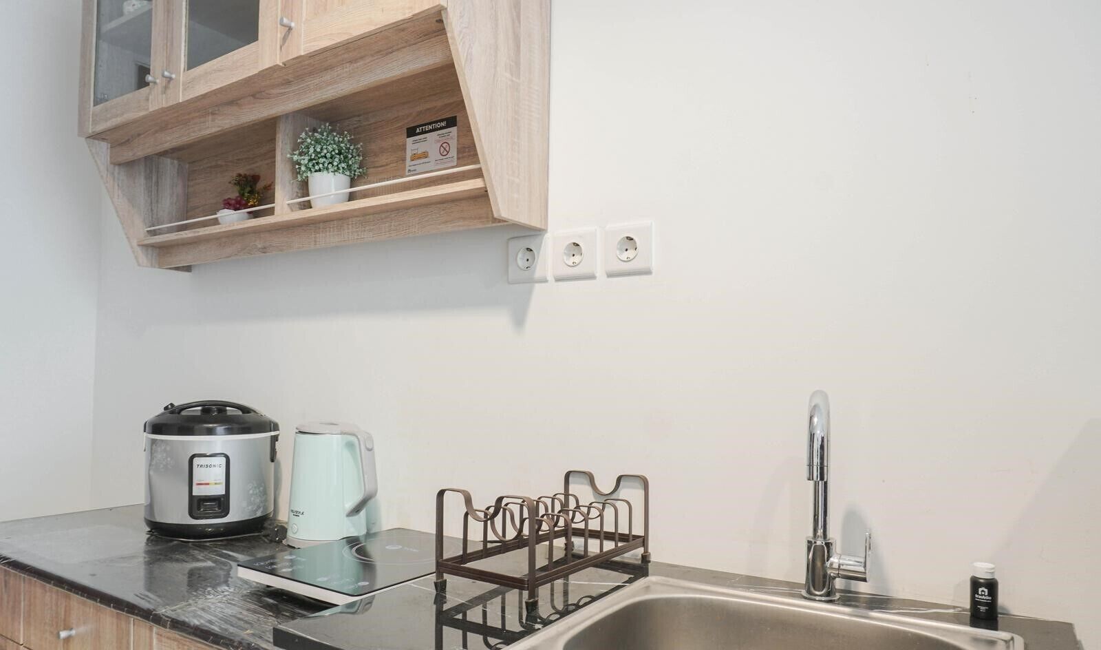 Una cocina con un lavamanos de acero inoxidable, una tetera, una cocedora de arroz y un armario con una planta.