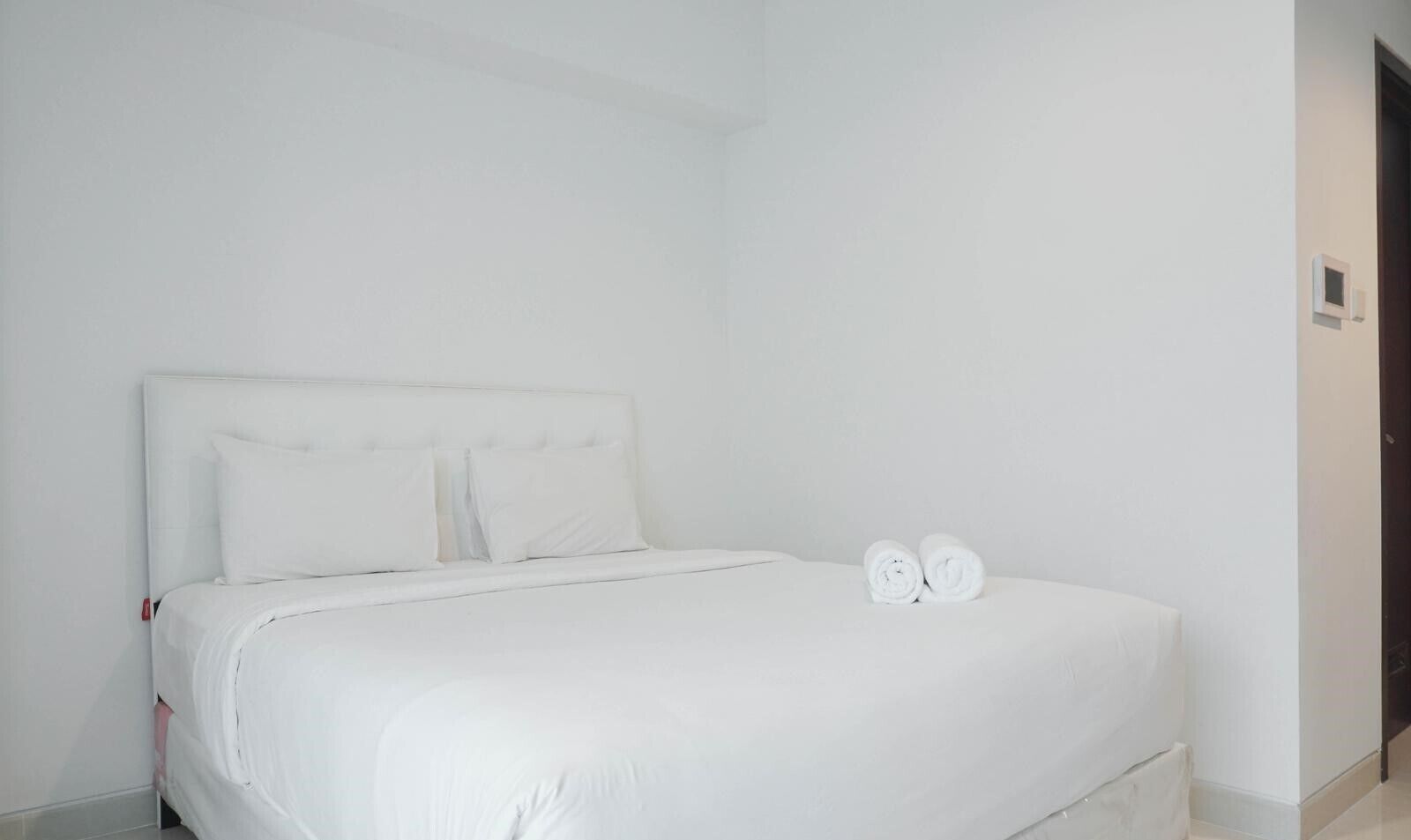 A white bed with two rolled towels, a white headboard, and a plain wall.