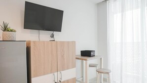 A modern kitchen with a flat-screen TV mounted above a wooden cabinet, a small table with a black storage box, and a white chair. - Scenic And Comfy Studio West Vista Apartment (Jakarta)