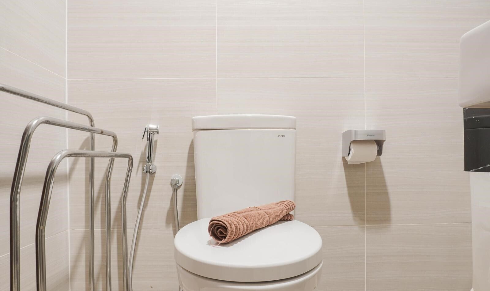 A bathroom with a white toilet, a towel, a handrail, and a paper towel dispenser.
