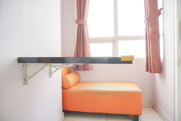 Una habitación pequeña e iluminada, con un banco naranja, un estante de madera y una ventana con cortinas.