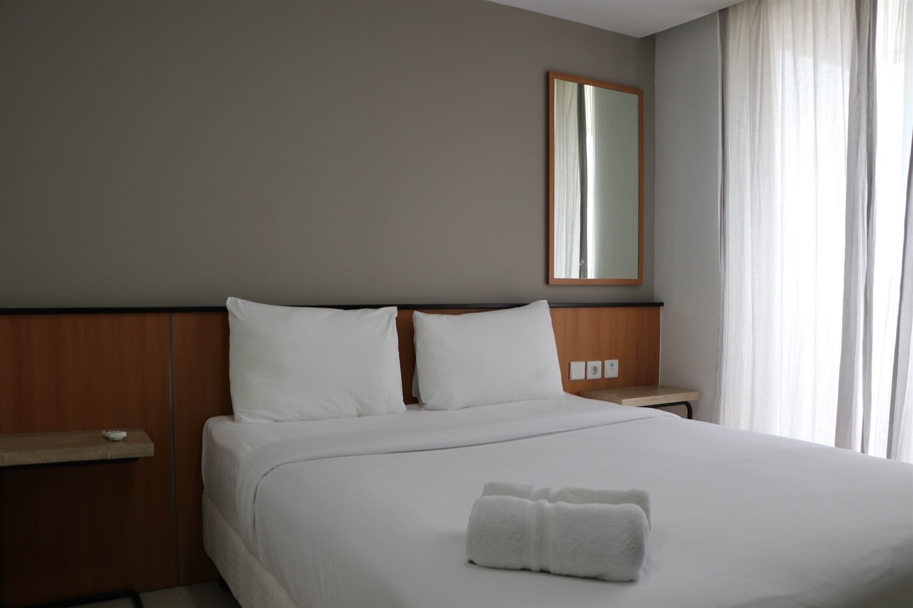 Una habitación de hotel con una cama impecable, un cabecero de madera, una mesita de noche con una lámpara y un espejo grande en la pared.