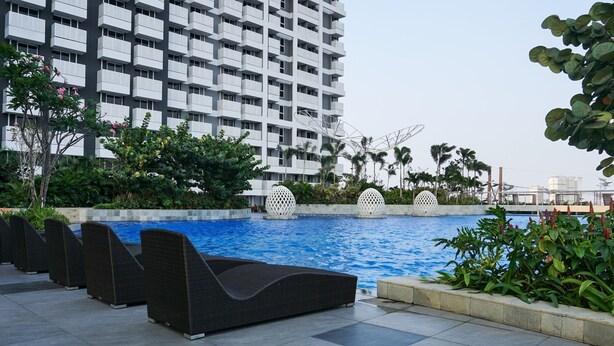 Un hotel moderno con piscina en la azotea, rodeado de exuberante vegetación y un rascacielos al fondo.