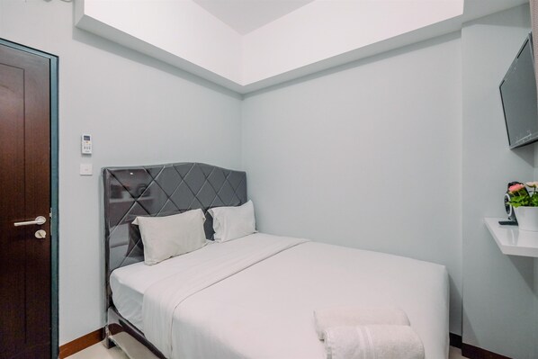 Una cama bien tendida con cabecera gris, una mesita de noche blanca con una planta y una puerta a la izquierda.