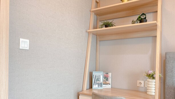 Uma secretária no canto, com cadeira, uma estante com plantas, um quadro emoldurado e um vaso com flores.