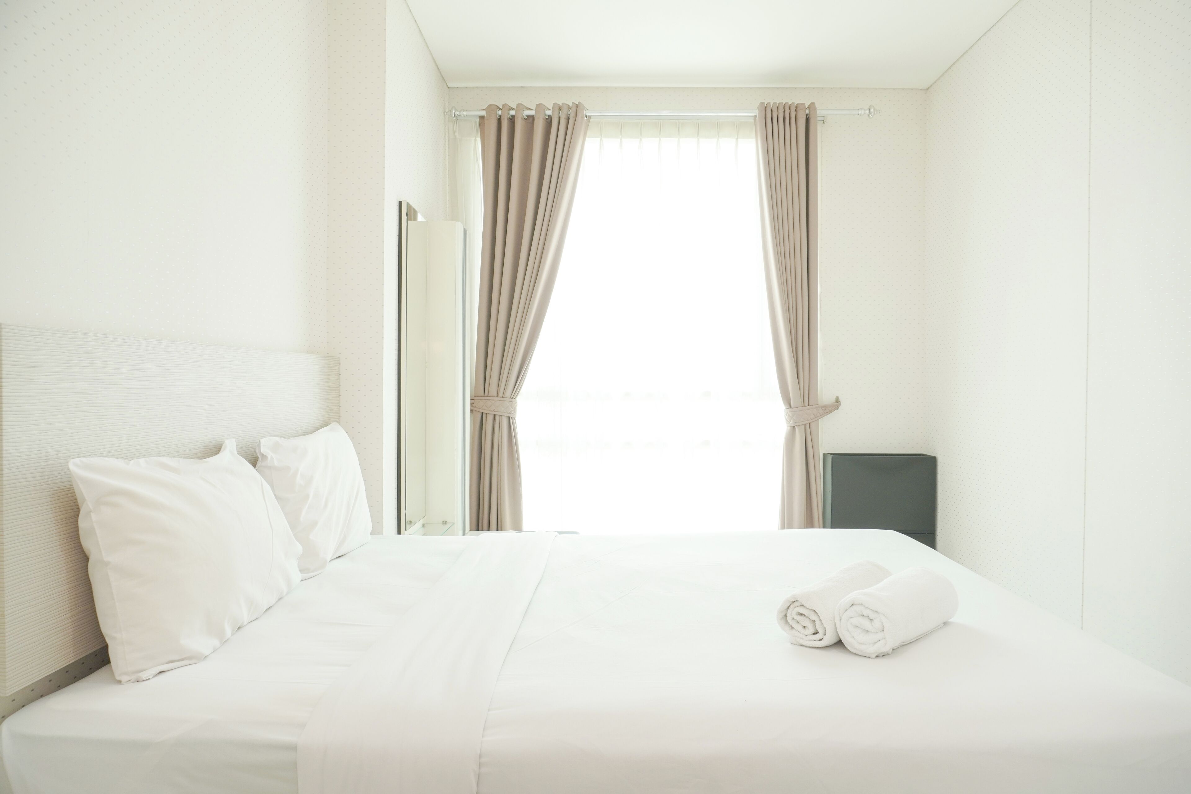 A hotel room with a neatly made bed, white linens, and a pair of rolled towels on the bed.