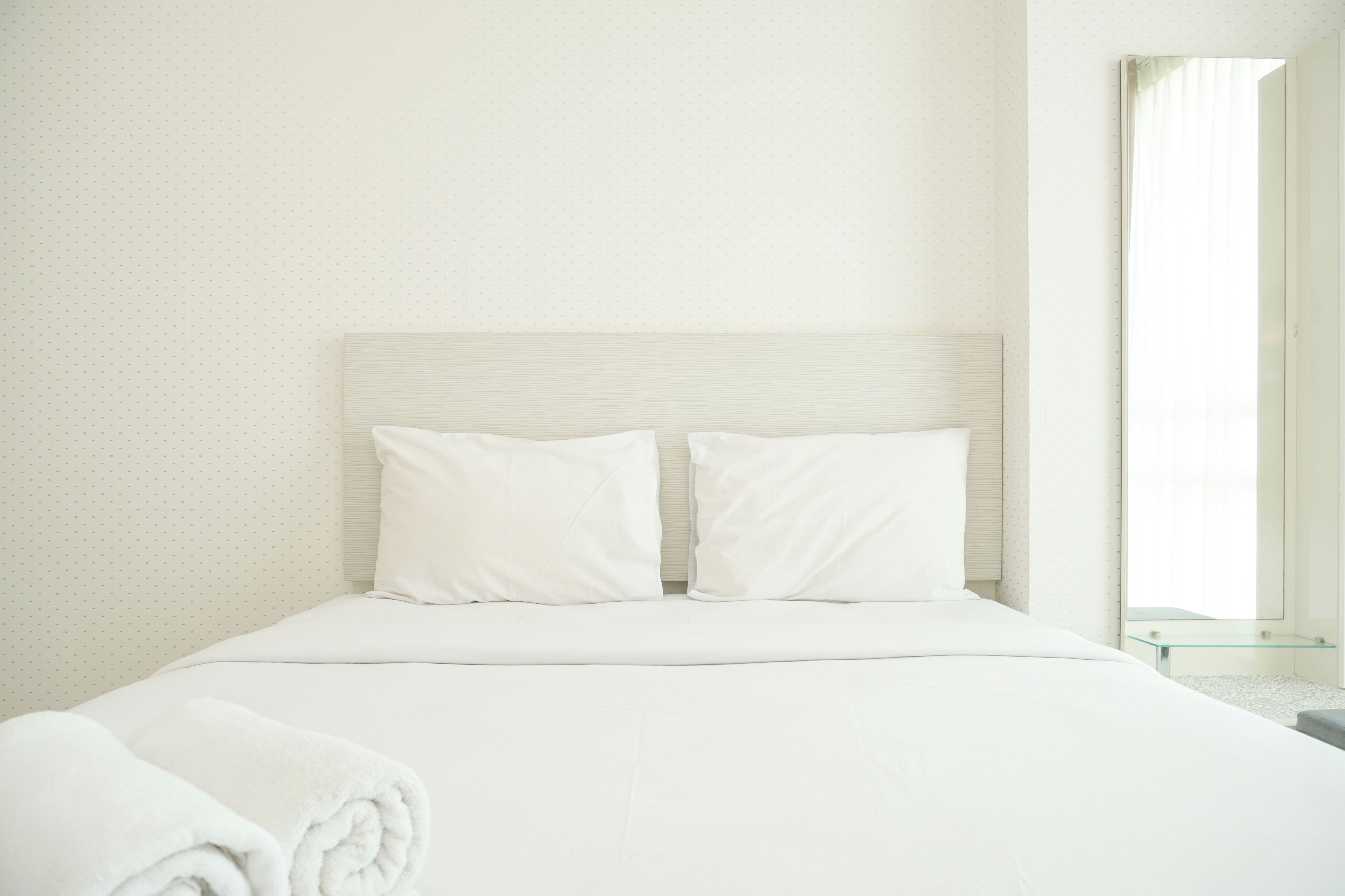 Una cama bien tendida con sábanas y almohadas blancas, una mesita de noche de cristal y una ventana con cortinas.