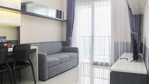 A modern, well-lit room with a grey sofa, a desk with a computer, and a chair. There is a large window with sheer curtains and a view of the outside. - Comfort And Cozy Living 1Br At Signature Park Grande Apartment (Jakarta)