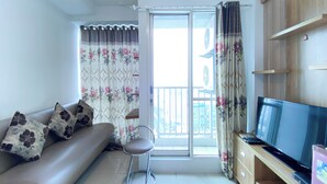 A living room with a sofa, a chair, a TV, and a balcony with curtains. - Good Deal And Homey 1Br Tifolia Apartment (Jakarta)