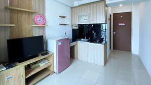 A compact kitchen with a refrigerator, microwave, and stove. There is a television on a wooden stand and a pink clock on the wall. - Good Deal And Homey 1Br Tifolia Apartment (Jakarta)