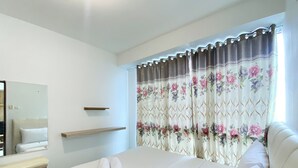 A bedroom with a bed, a bedside table, a mirror, and a window with floral curtains. - Good Deal And Homey 1Br Tifolia Apartment (Jakarta)