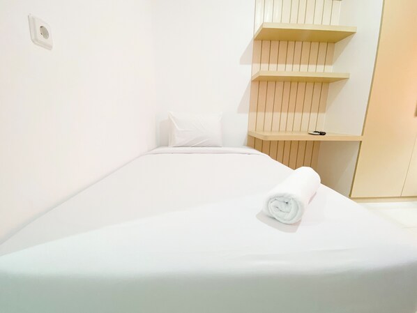 A neatly made bed with a rolled towel, a white pillow, and a small shelf with a book and a pair of shoes.