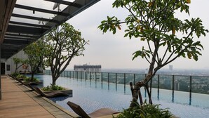 An infinity pool with a view of a cityscape, surrounded by lounge chairs and potted plants. - Comfy Stay 2Br Apartment At Arumaya Residence (Jakarta)