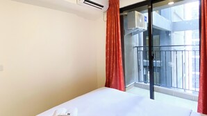 Ένα δωμάτιο ξενοδοχείου με ένα κρεβάτι, μια μονάδα κλιματισμού, ένα παράθυρο με κόκκινες κουρτίνες και θέα στο εξωτερικό μπαλκόνι.