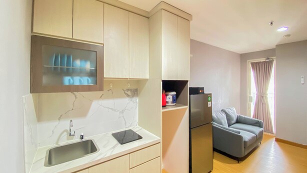 A modern kitchen with a built-in oven, a sink, and a refrigerator. There is a grey sofa and a window with curtains in the living area. - Comfort Living 2Br At Cinere Bellevue Suites Apartment (Depok)