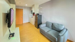 A modern living room with a grey sofa, a flat-screen TV mounted on the wall, a kitchen area with a refrigerator and cabinets, and a wooden door. - Comfort Living 2Br At Cinere Bellevue Suites Apartment (Depok)