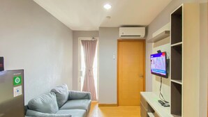 A living room with a grey sofa, a flat-screen TV mounted on a white cabinet, and a wooden door. - Comfort Living 2Br At Cinere Bellevue Suites Apartment (Depok)