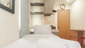 A neatly made bed with white linens and a rolled towel, a wooden door, a small desk with a chair, and a clock on the wall. - Good Deal And Restful Studio Apartment B Residence (Pagedangan)