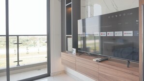 A modern living room with a large flat-screen TV mounted on a wooden cabinet, a remote control on the cabinet, and a view of a cityscape through a large window. - Comfortable And Good Place Studio At Sky House Bsd Apartment (Pagedangan)