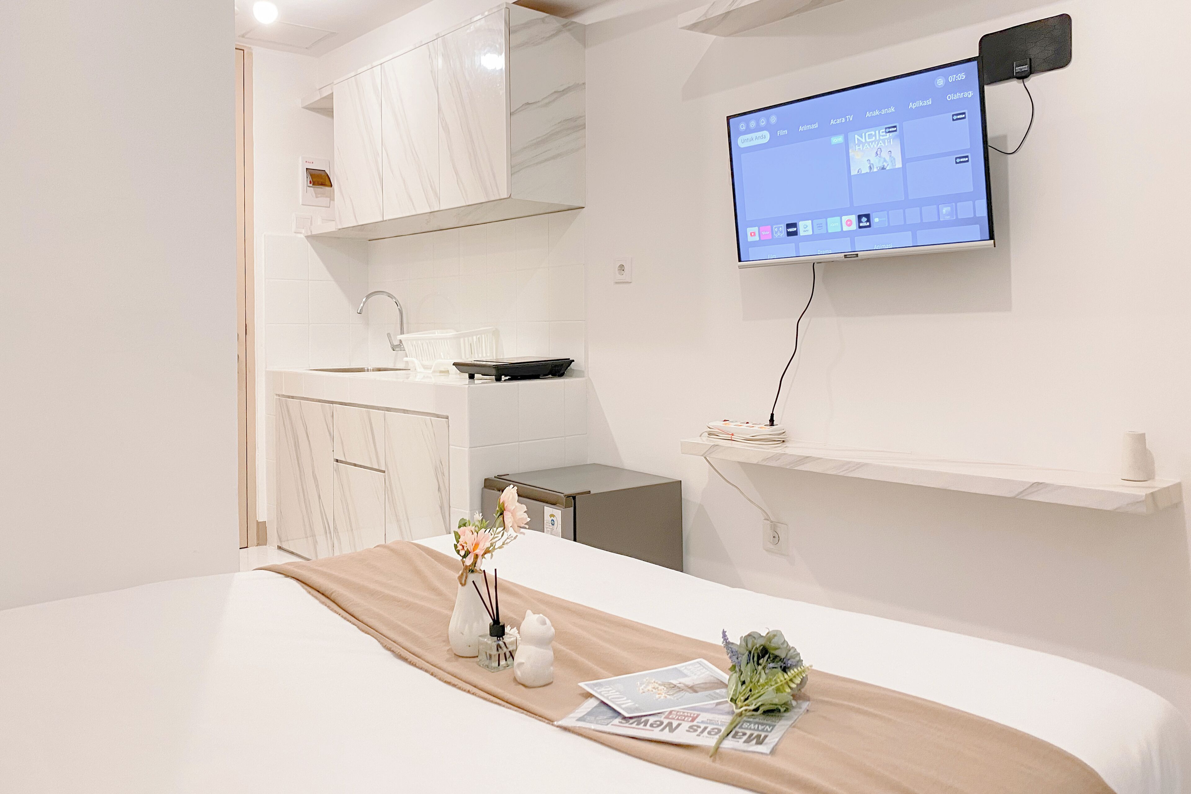 A modern hotel room with a bed, a TV mounted on the wall, a small kitchen area, and a shelf with a vase.