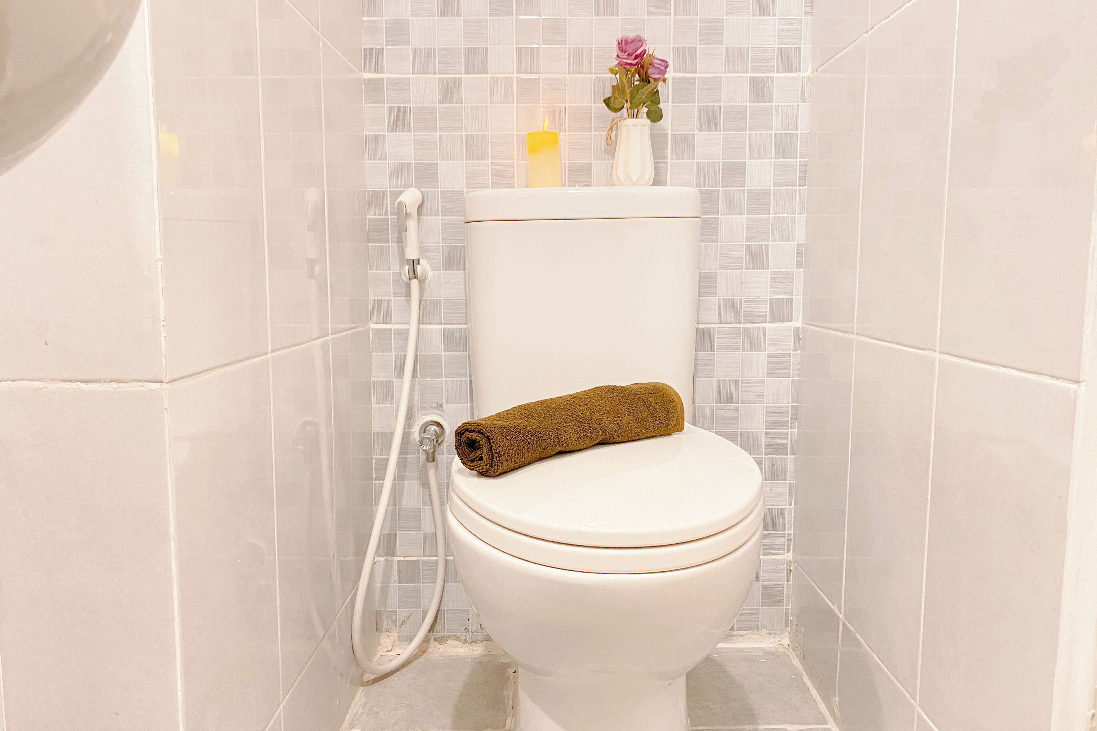 A toilet with a rolled towel on the seat, a small vase with flowers, and a candle on the tank.