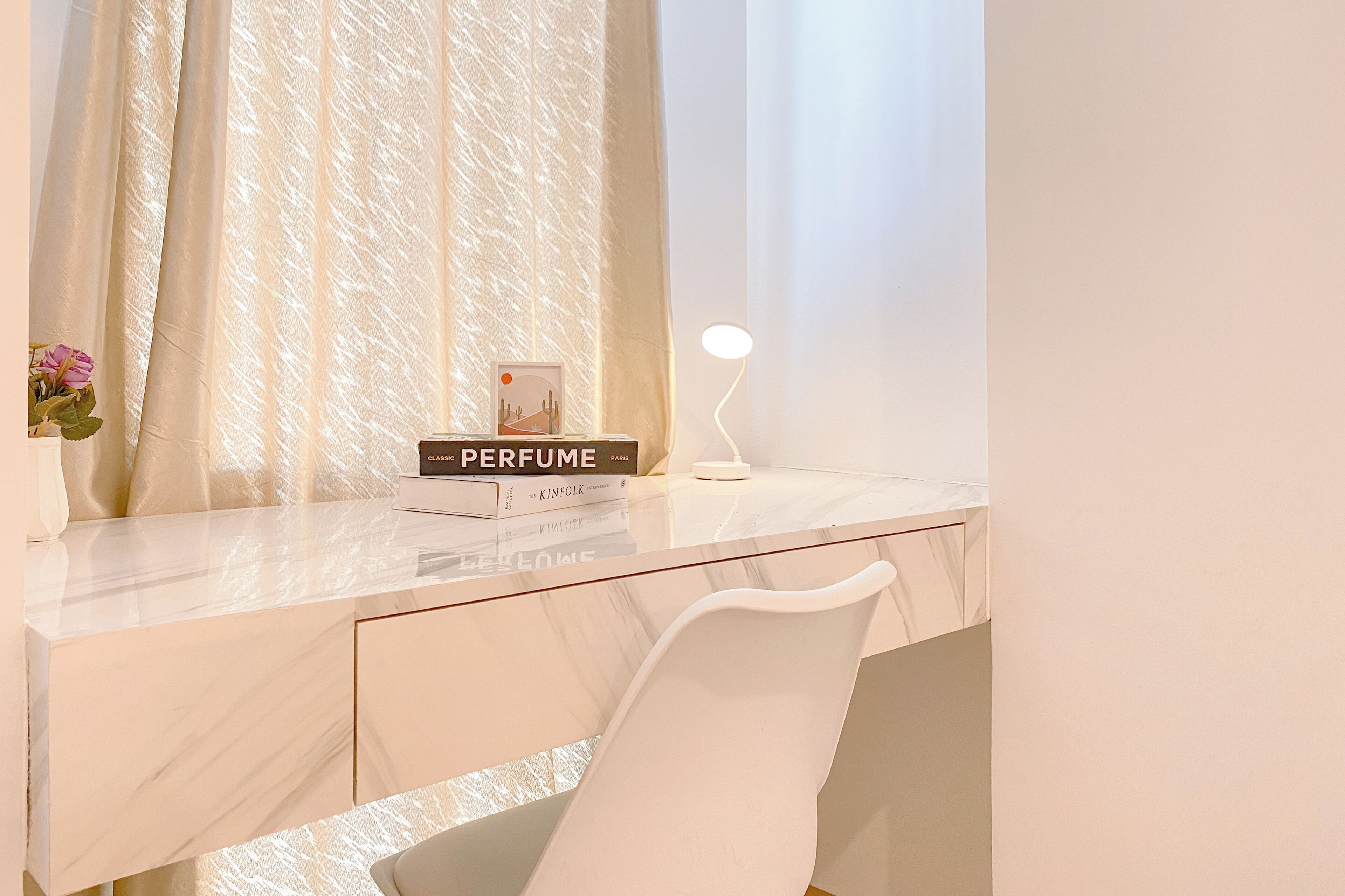 Un escritorio moderno con cubierta de mármol, una silla blanca, una lámpara pequeña y una pila de libros, incluyendo 'Perfume' de Patrick Süskind.