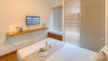 Ein Hotelzimmer mit einem Bett, einem an der Wand montierten Fernseher, einem Fenster mit Jalousien und einem Holzboden mit einer Vase und einer kleinen Pflanze.