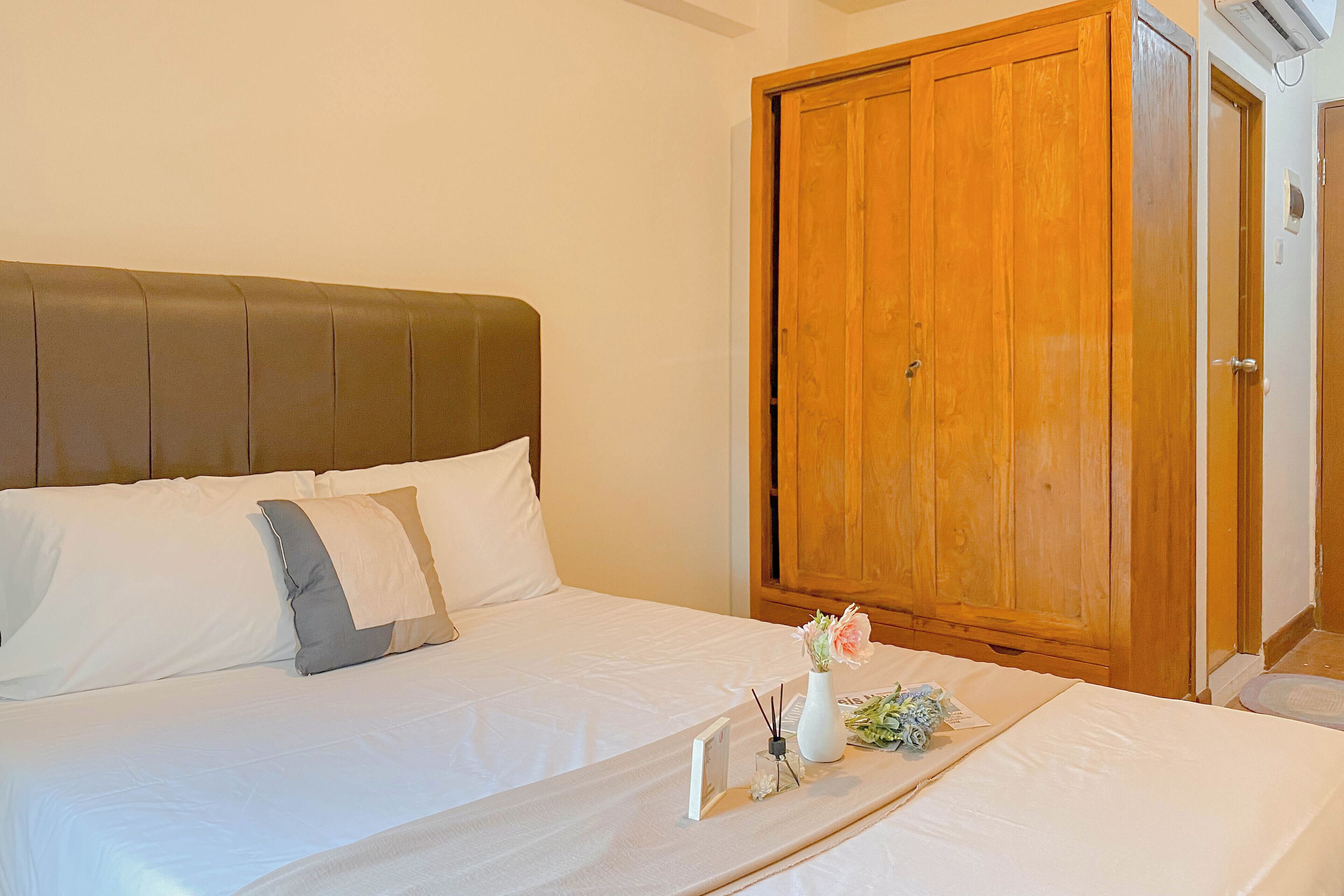 Ein ordentlich bezogenes Bett mit weißen Leinen und einem braunen Kopfteil, ein Holzschrank und ein kleiner Nachttisch mit Blumen in einer Vase.