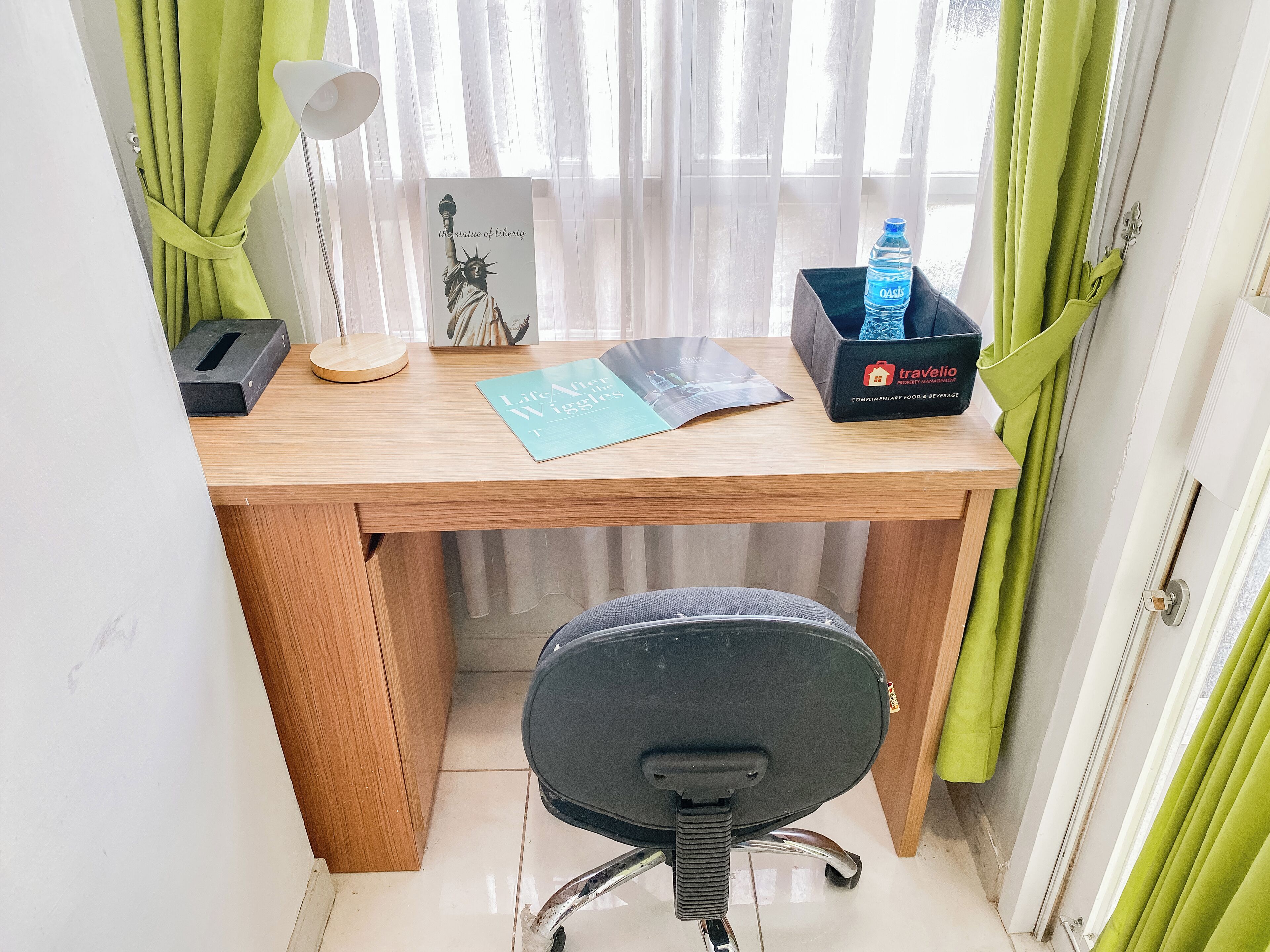 A small, neatly organized workspace with a wooden desk, a black office chair, a lamp, a framed picture of the Statue of Liberty, a water bottle, and a box labeled 'Traveling'.