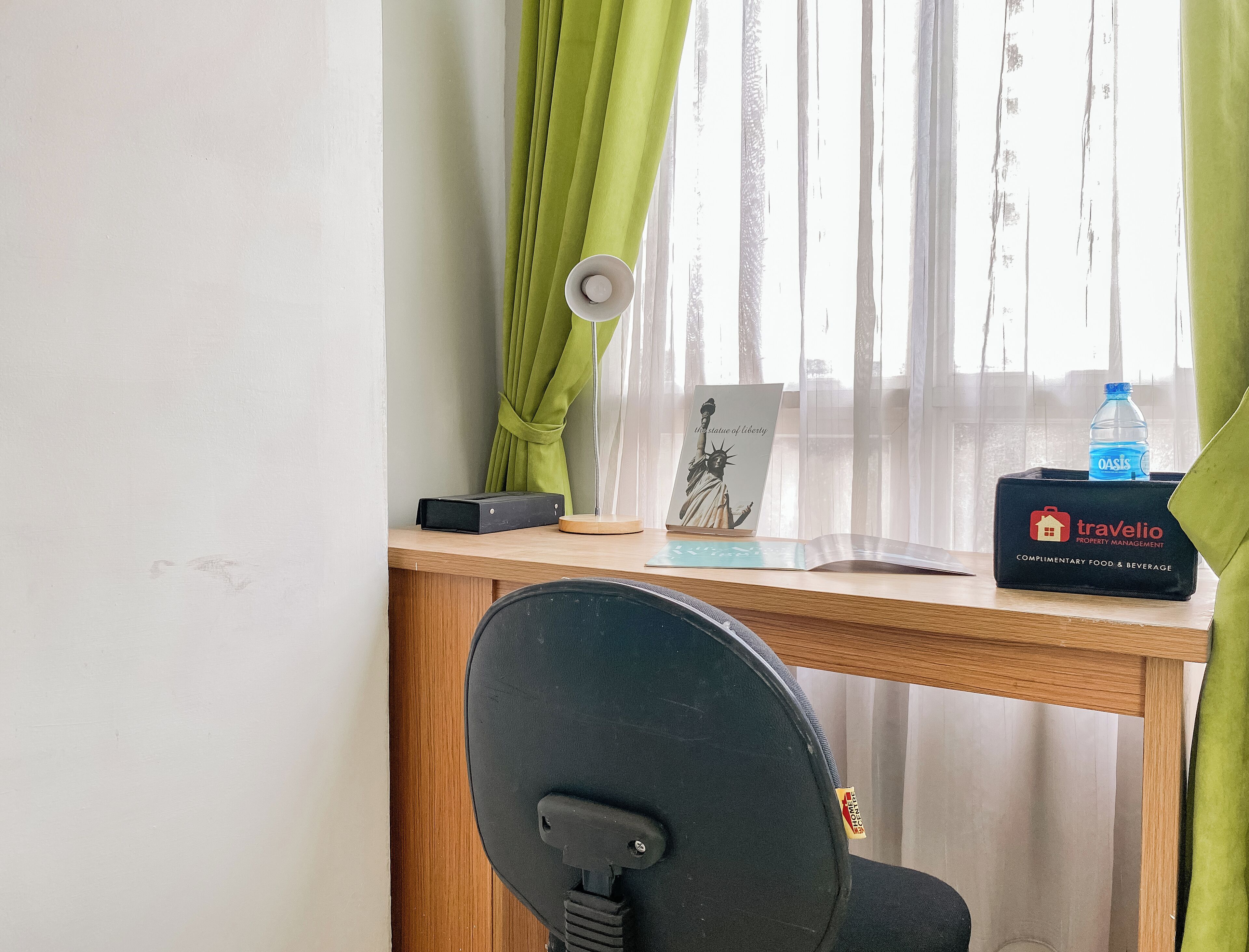 A hotel room with a wooden desk, a black office chair, a lamp, a framed picture of the Statue of Liberty, a water bottle, and a box labeled 'Travelio Property Management'.